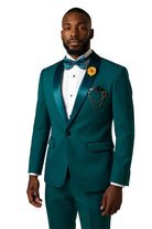 A man wearing a teal emerald green tuxedo with satin peak lapels, a white pinstripe shirt, a matching teal bow tie, and a yellow flower boutonniere, standing against a light gray studio background.