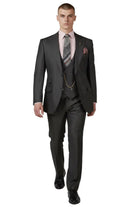 A man modeling a dark charcoal gray three-piece suit with a light pink dress shirt, a patterned gray tie, and a pocket square, standing against a light gray studio background.