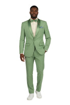 A man modeling a light sage green two-piece suit with satin shawl lapels, a matching green bow tie, a cream dress shirt, and white sneakers against a plain white studio background.