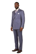 A man modeling a double-breasted suit in a dusty lavender-blue color, paired with a white shirt, a patterned purple tie, a white pocket square, and brown leather dress shoes, set against a plain white studio background.