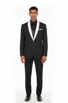 A man modeling a black tuxedo with white satin lapels, a black shirt, a black bow tie, and a white pocket square, shot against a plain white studio background.