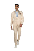 A man modeling a light beige two-piece suit with satin peak lapels, a light blue dress shirt, a matching beige bow tie, a floral boutonniere, and white sneakers, set against a plain white studio background.