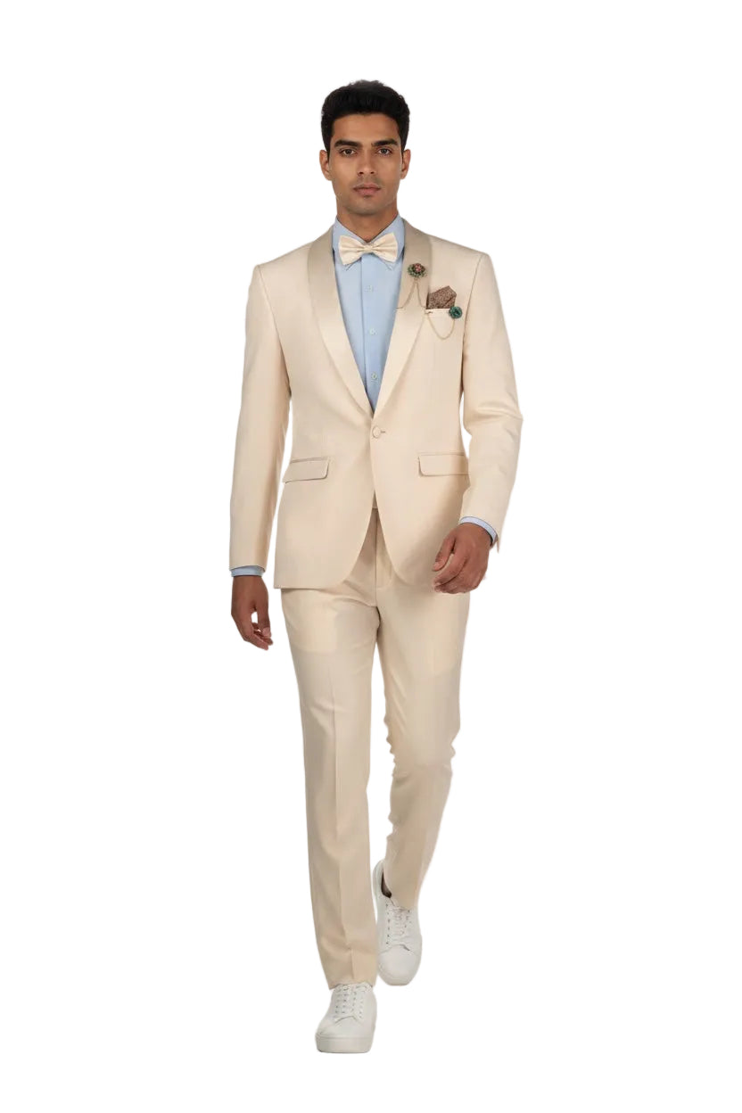 A man modeling a light beige two-piece suit with satin peak lapels, a light blue dress shirt, a matching beige bow tie, a floral boutonniere, and white sneakers, set against a plain white studio background.