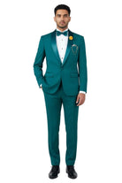 A man modeling a teal green tuxedo with satin lapels, a white dress shirt, a matching green bow tie, and a yellow flower boutonniere, standing against a light gray studio background.