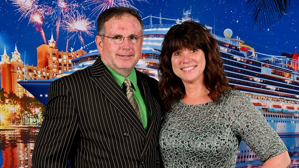 A man in a black pinstripe suit with a green shirt and tie stands next to a woman in a patterned gray-green top, posing in front of a cruise ship and fireworks backdrop.