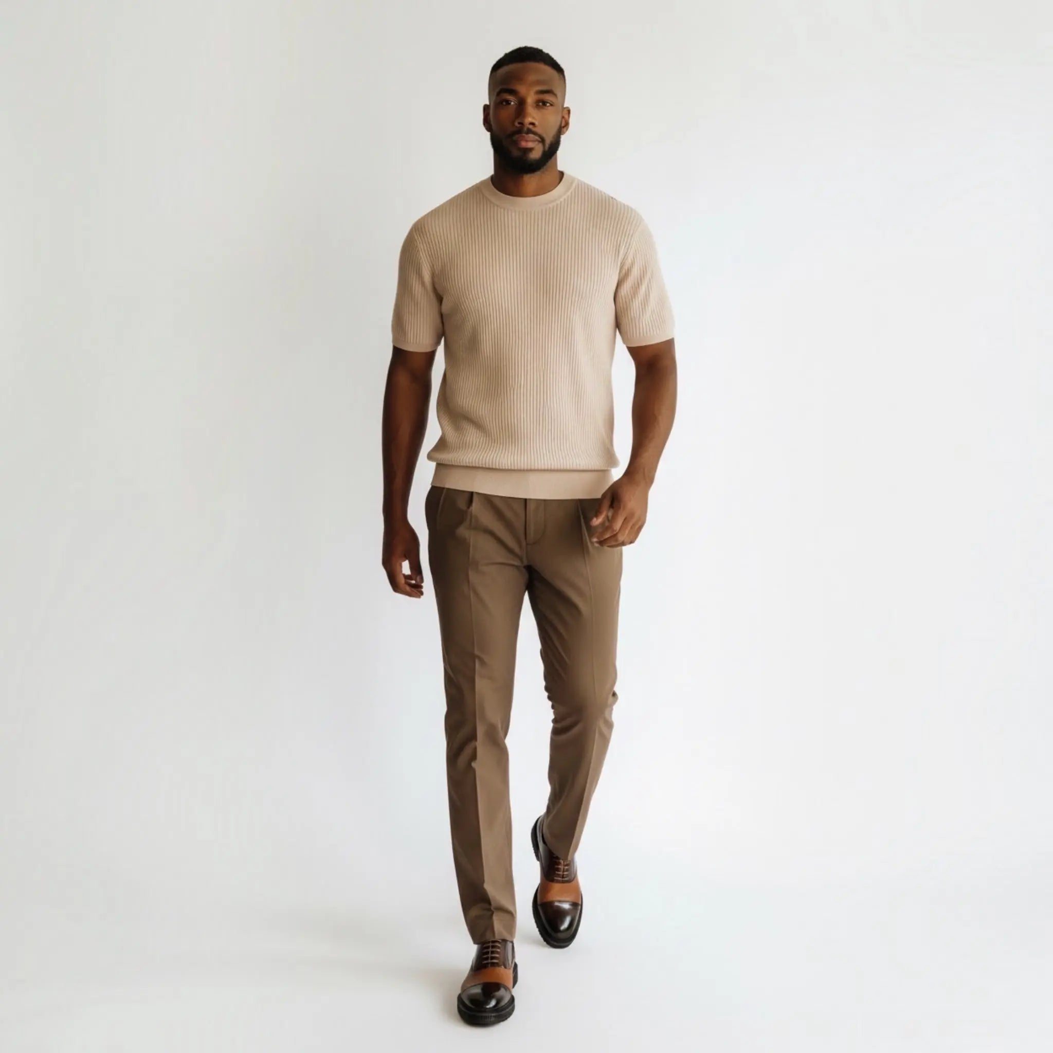 A man modeling a light beige short-sleeve knit sweater and brown trousers against a plain white studio background.
