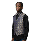 A young boy modeling a formal outfit featuring a patterned waistcoat, a black dress shirt, a matching patterned bow tie, and blue jeans, set against a plain light gray background.