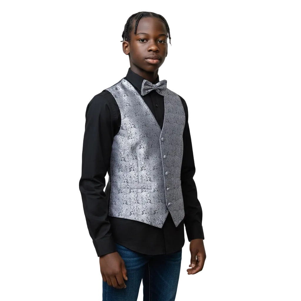 A young boy modeling a formal outfit consisting of a black long-sleeve dress shirt, a silver patterned vest, a matching silver bow tie, and blue jeans, set against a plain light gray studio background.