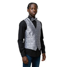 A young boy modeling a formal outfit consisting of a black long-sleeve dress shirt, a silver patterned vest, a matching silver bow tie, and blue jeans, set against a plain light gray studio background.